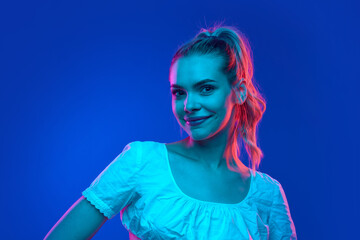 Young attractive blonde woman looking at camera with light smile in neon light against gradient studio background. Trends. Concept of human emotions, carefree, beauty and youth, fashion and style. Ad