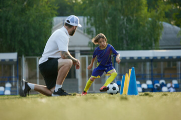 Obraz premium Young athlete, boy, child in uniform in motion, improving dribbling skills under watchful eye of professional coach. Outdoor training session. Concept of sport, childhood, education, active lifestyle
