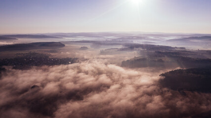 Aerial view of rolling hills covered with dense mist and a bright sun in the morning sky