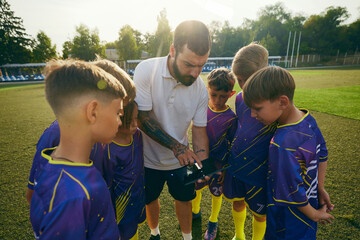 Young athletes, boys listening with attention to their coach explaining game plan with digital tablet. Sunny evening game. Concept of sport, childhood, education, achievement, active lifestyle