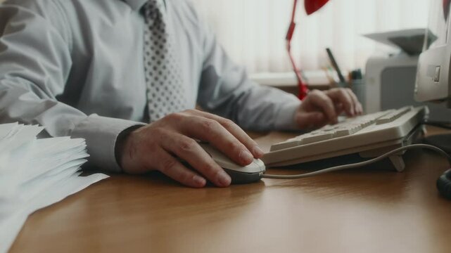Cropped shot of unrecognizable clerical worker in shirt and tie sitting at wooden table next to pile of papers, using old wired mouse while working on retro computer indoors, nostalgic 80s