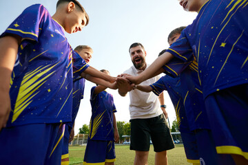 Fototapeta premium Young soccer players and their coach unite with hands in, showing team spirit and enthusiasm. Motivation before game. Concept of sport, childhood, education, achievement, active lifestyle