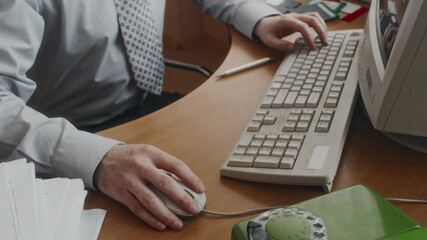 High angle shot of hands of faceless clerical worker in shirt and tie working on vintage computer, using old keyboard and wired mouse at wooden desk, next to documents and rotary phone in 80s office
