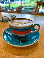 A close-up of a creamy cappuccino served in a vibrant striped cup with a matching saucer and spoon, set on a wooden table in a cozy cafe, with sunglasses and a scenic view in the background