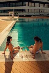 Two women relaxing by the poolside with glasses of chilled drinks. The sun shines brightly,...