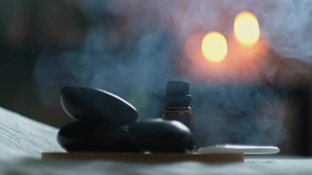 Spa salon equipment. Close up shot of massage hot stones with bottle of massage oil on towels with flaming candles and smoke from incense sticks or incense background. wellness, spa concept 4k footage