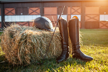 equestrian horse riding equipment with helmet, leather boots and stick on grass and hay bale, barn...