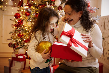Mother and daughter unwrapping Christmas gift in festive holiday setting with decorated tree