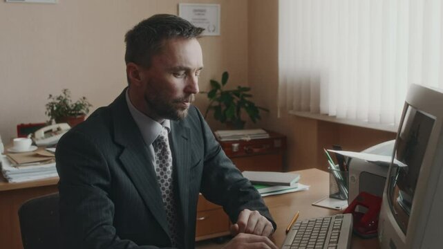 Side medium shot of experienced bearded Caucasian office worker giving advice about documents to diverse colleague and continuing working on outdated computer in 90s retro office