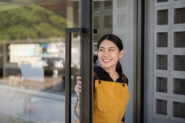 Young Barista in Yellow Apron Smiling While Opening a Glass Door of a Modern Cafe