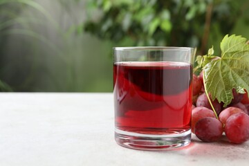 Tasty grape juice in glass, leaf and berries on light table against blurred green background, closeup. Space for text