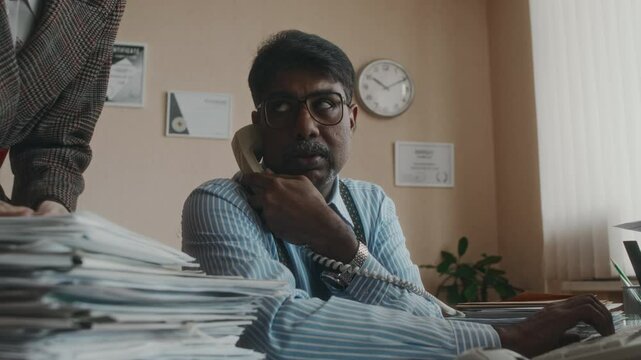 Side shot of hardworking diverse mature office worker in formalwear answering call on wireline phone and working on old computer, colleague adding documents to pile of papers to verify in 90s
