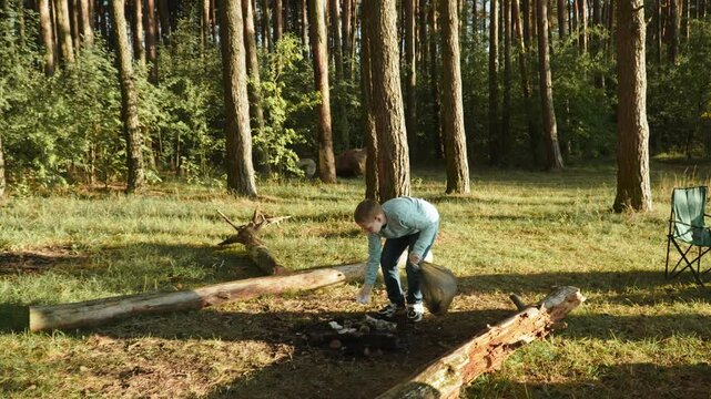 Child collects thrown garbage in bag in forest from fire pit in sunny weather. Boy volunteers and helps clean up campsite after holiday. Good behavior and education of younger generation.