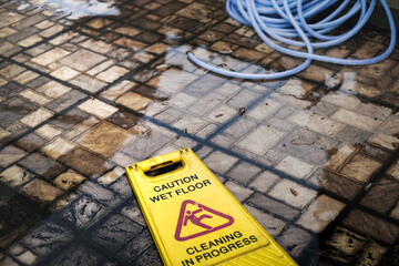 Yellow Caution Wet Floor Cleaning in Progress Sign and Gardening Hose Fell on Dirty Mossy Wet and Flooded Outdoor Floor