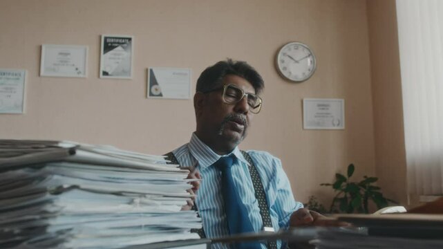 Medium shot of male Biracial clerk in years taking papers from stack, sitting at desk full of documents, looking tired, stretching arms and putting head on arms, having headache from work in office