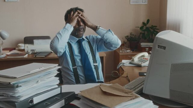 Medium shot of mature Biracial employee in formalwear working with myriads of documents, taking off glasses and putting arms around head, having severe headache from monotonous work in retro office