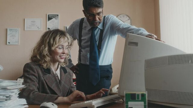Medium shot of mature Biracial boss in formal wear coming up to young female Caucasian apprentice to check her working on old computer in nostalgic 80s office