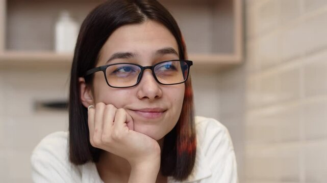 Young woman in glasses thoughtfully resting her chin on her hand, remember something pleasant and smiling with satisfaction at home close up
