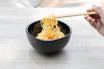 hands using chopsticks, pick up noodles in a black bowl with instant noodles on wooden table.