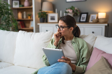 Attractive woman, freelancer holding and using digital tablet, sitting at cozy sofa in living room. Successful businesswoman text message in home. Online technology concept, remote job and work.