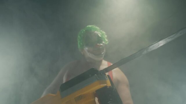 Scary man in the horror clown mask with green hair, standing in suspenders in the dark studio with flashing lights and threatening with the chainsaw close up