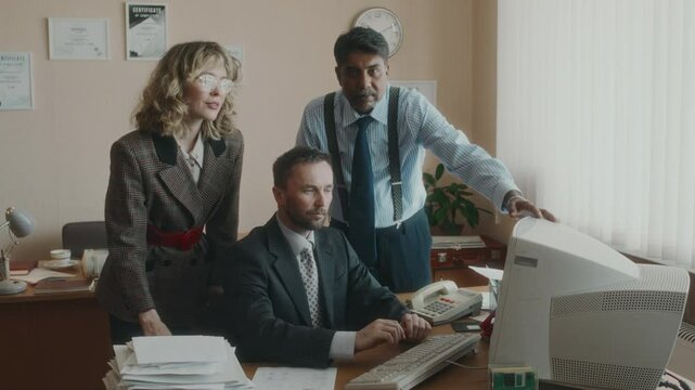 Medium long shot of group portrait of diverse colleagues in formal wear gathered around clerical worker working on outdated computer and looking up at camera together, retro office in 90s