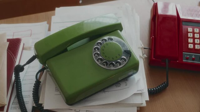 No people top view of green olive colored rotary telephone on ream of documents on wooden table next to bright red retro landline phone in 90s office