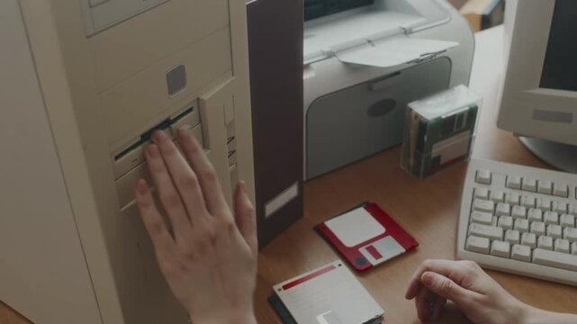 High angle shot of hands of anonymous clerical worker choosing green floppy diskette and inserting it into outdated computer while working at desk in office in 90s