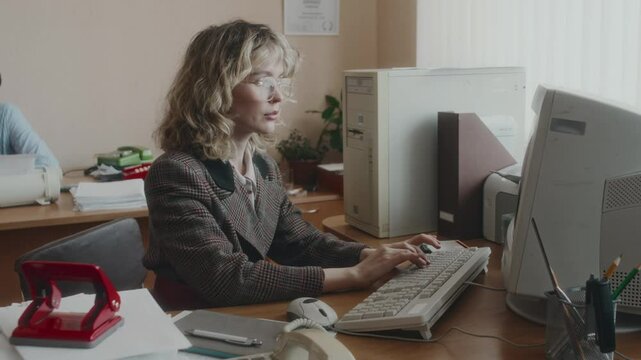 Side shot of female Caucasian clerical worker with blonde wavy hair wearing checkered jacket and eyewear, working at desk on large monitor and inserting floppy disc into computer in office, 90s