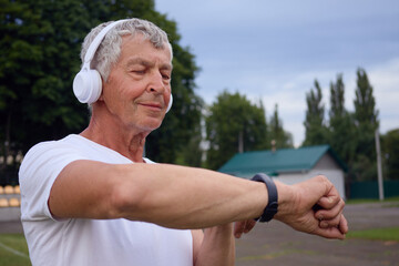 Technology for fitness routines outside. Middle-aged gentleman in white shirt examining his heart rate or burning calories on his smartwatch standing at the stadium using wireless headphones