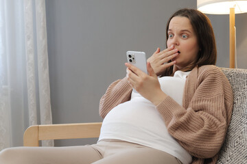 Surprised astonished pregnant woman using smartphone for browsing web pages reading shocked...