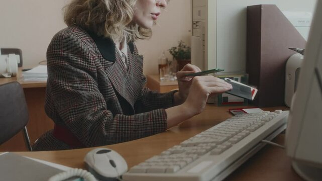 Side tilt shot of pretty blonde Caucasian employee in checkered jacket and square glasses picking floppy disc and inserting it into old retro computer during workday in office in 90s