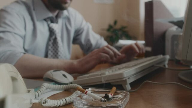 Cropped shot of two smouldering cigarettes in ashtray on wooden office desk, anonymous office worker working on computer in background, 90s