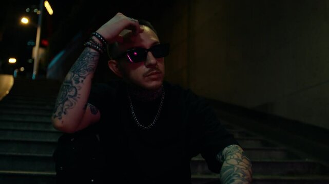 Cool guy with tattoos and sunglasses, hanging out in underground subway station, green lights.