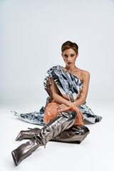 Woman in shimmering, recycled outfit sits on white background, silver boots adding futuristic touch