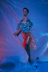 A woman wearing a silver jacket and red pants stands surrounded by blue and pink plastic.