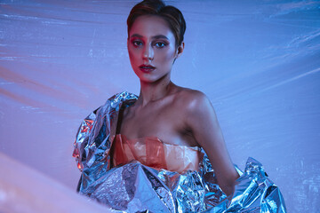 A woman poses in a silver foil dress, bathed in blue light.
