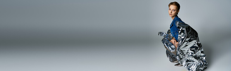 Woman sits on gray background, draped in shimmering silver, with reflective gaze