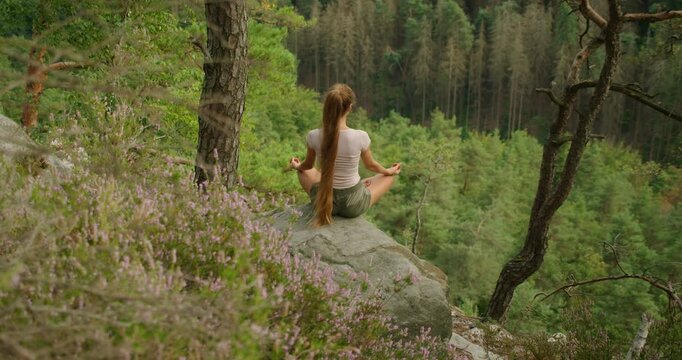 Woman in a meditative pose on a cliffside overlooking a vast green forest in summer day.