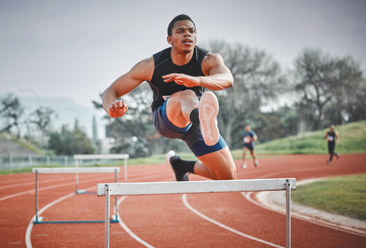 Hurdles, fitness and man running on track at stadium training for race, competition or marathon. Energy, jump and male athlete sprint by equipment for cardio workout or exercise at sports arena.