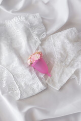 Pink reusable menstrual cup lies on white lace underwear on white sheet, concept of feminine intimate hygiene and zero waste. There are small flowers in the menstrual cup. Minimalism. Copy space.