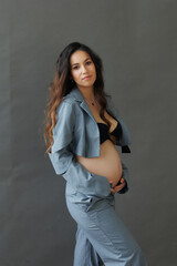 A beautiful pregnant brunette woman in a cropped gray stylish suit on a gray paper background in the studio. A pretty girl with a bare stomach in a fashionable suit is sitting barefoot on a gray backg