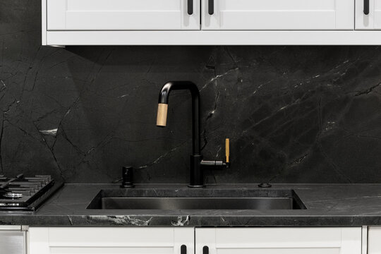 A kitchen sink detail with a modern black and gold faucet, with a black stone countertop and backsplash, and white cabinets.