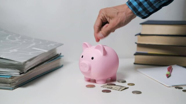 Hand drops a coin into a pink piggy bank on a white table, surrounded by scattered coins and a crumpled dollar bill. Books and a notebook with a pen sit in the background