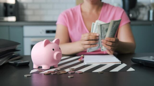 Young woman counting banknotes while sitting at kitchen table, a piggy bank and loose change are scattered in the foreground, selective focus. Personal finances, budgeting and savings concept