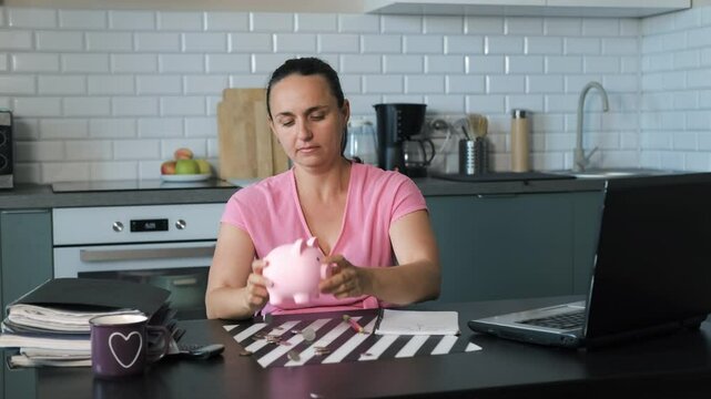 Young woman shaking money out of pink piggy bank onto a kitchen table with coins scattered around. Concept of budgeting and managing personal finances