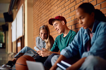 Happy teenagers using smart phone while relaxing in hallway at high school.