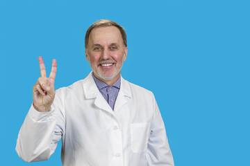 Happy male senior medic in white coat shows victory gesture sign. Isolated on blue.