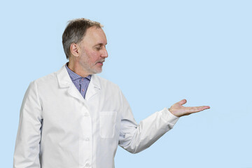 Portrait of senior doctor advertising your product on his hand. Physician with copy space. Isolated on pastel blue.