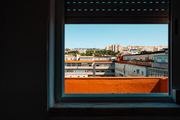 View through a window frame of a modern residential buildings with rooftops and blue sky. Urban landscape, architectural lines, highlighting the contrast between indoor darkness and outdoor brightness